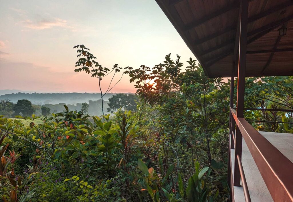 Sunset view from a wooden balcony overlooking lush tropical forest and distant hills.