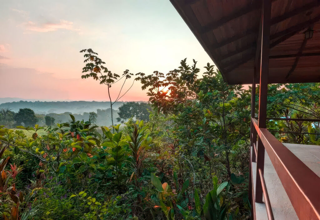 Sunset view from a wooden balcony overlooking lush tropical forest and distant hills.