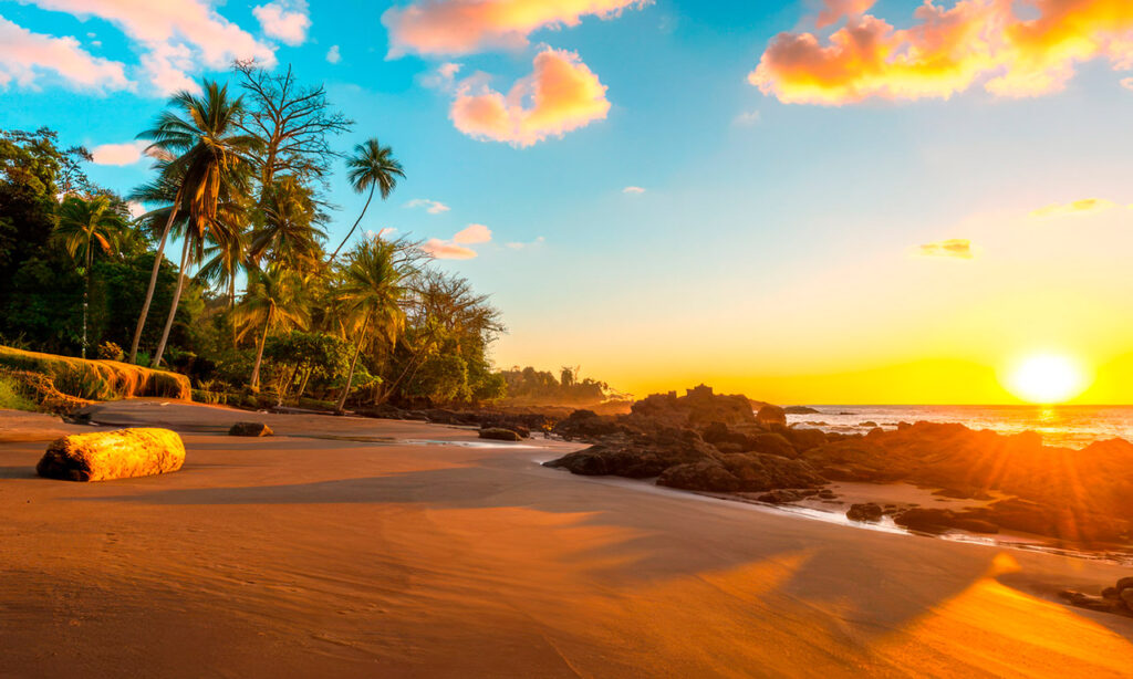 Vibrant sunset over a pristine beach in Corcovado National Park, Costa Rica, with palm trees, rock formations, and golden sand reflecting the sun's rays.